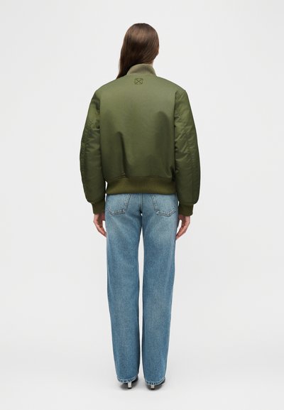 Άτομο με μακριά καστανά μαλλιά που φοράει μπουφάν bomber σε ελιά πράσινο και άνετο μπλε τζιν, στέκεται μπροστά σε ένα απλό λευκό φόντο.