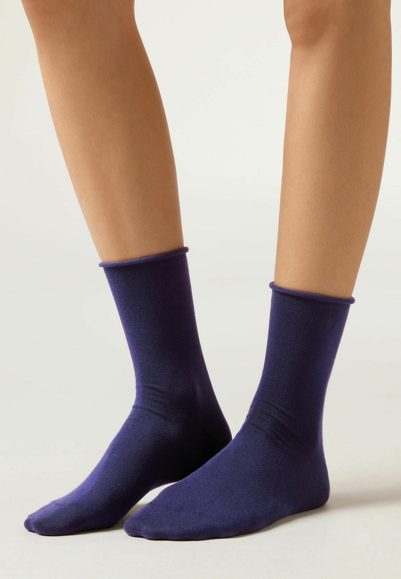 Gambe che indossano calze semplici di un viola scuro a metà polpaccio su uno sfondo neutro chiaro.