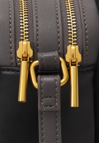 Détail d'un sac en cuir gris avec des fermetures à glissière doubles en ton doré, présentant un fermoir en métal doré et une bandoulière en cuir assortie, mettant en valeur la texture et le design.