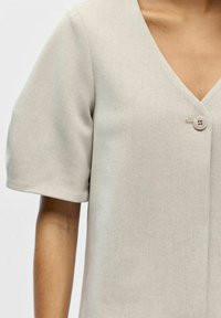 Vêtement gris clair à manches courtes avec un col en V et un tissu texturé. Dispose d'une fermeture à un bouton au niveau de la poitrine.