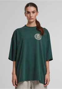 Veľká tmavozelená tričko z bavlny s okrúhlym výstrihom, krátkymi rukávmi a kruhovým logom potlačeným na ľavej hrudi.