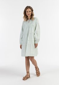 Helles blaues, gemustertes Kleid mit einem Ausschnitt in Schlüssellochform, langen Ärmeln und gestuften Design. Kombiniert mit beigen Riemchensandalen.