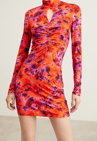 Robe mini à manches longues en tissu orange vif avec des motifs floraux violets et noirs, présentant un décolleté en forme de clé et des détails froncés.