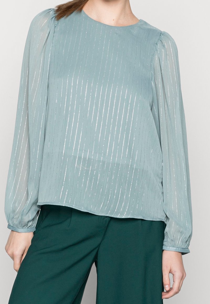 Blusa a maniche lunghe blu chiaro con strisce metalliche verticali, scollatura rotonda e maniche svasate. Abbinata a pantaloni verde scuro.