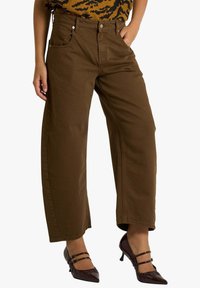 Pantaloni larghi in cotone marrone con due tasche frontali, dettagli in metallo e vita alta, abbinati a décolleté bordeaux con punta appuntita.