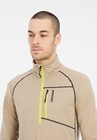 Bežinis fleece puloveris su ketvirčio užtrauktuku, juodomis siuvimo detalėmis ir ryškiai geltonu užtrauktuku. Yra užtraukiama krūtinės kišenė su logotipu.