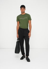 Grønn bomull t-skjorte med hvit tekst, svarte bukser med elastisk midjebånd, svarte sko og en svart tote bag, satt mot en ensfarget bakgrunn.