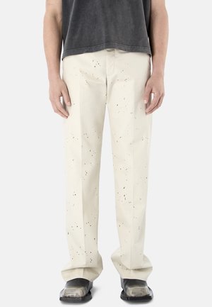 Persona che indossa pantaloni color avorio con motivo a macchie nere, abbinati a scarpe nere con punta quadrata e una camicia corta grigio scuro.