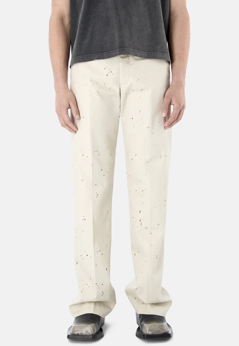 Persona con pantalones color marfil con patrón de manchas negras, combinados con zapatos negros de punta cuadrada y una camisa corta gris oscuro.