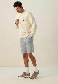 Sweatshirt crème avec un design de poche géométrique, shorts rayés bleu et blanc, baskets grises et chaussettes blanches avec des rayures jaunes.