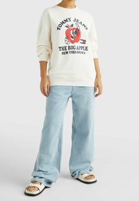 Felpa crema con stampa grafica di una mela rossa e testo "TOMMY JEANS THE BIG APPLE"; abbinata a jeans a gamba larga di un blu chiaro e sandali bianchi.