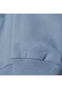 Pulover albastru deschis, cu manșete și tiv cu nervuri. Prezintă un logo circular embosat subtil pe mânecă. Textură moale, design lejer.