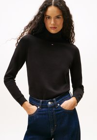 Donna con lunghi capelli ricci, che indossa un maglione a collo alto nero e jeans a vita alta blu scuro, con le mani nelle tasche, sfondo neutro.