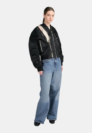 Donna in piedi che indossa una giacca nera oversize con zip sopra una canotta beige, jeans blu a gamba larga e scarpe nere su uno sfondo semplice.