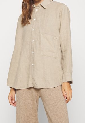 Kobieta ubrana w beżową, długorękawową koszulę zapinaną na guziki z kieszenią z przodu oraz jasnobrązowe, dziane spodnie na tle jednolitego tła.