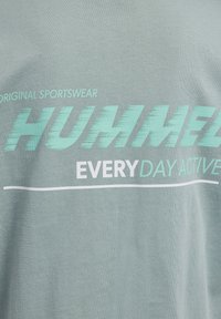 Szara tkanina z turkusowym i białym napisem "Original Sportswear HUMMEL Everyday Active" oraz białym podkreśleniem poniżej.
