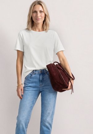 Lyshåret kvinde iført hvid t-shirt og blå jeans holder en stor mørkerød håndtaske mod en enkel lys baggrund.