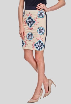 Mujer con una falda beige con patrones geométricos azules y rojos, combinada con zapatos de tacón color nude, de pie frente a un fondo liso.