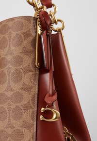 Sac à main en cuir marron à motifs avec un accent lisse rouge, des ferrures dorées et un charme décoratif. Dispose d'une bandoulière en chaîne pour plus de polyvalence.