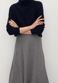 Persona vistiendo un suéter negro de cuello alto con los brazos cruzados y una falda midi texturizada gris, adornada con anillos de oro.