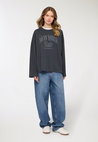 Felpa oversize grigia con grafica "NEW YORK State", abbinata a jeans denim blu larghi e sneakers bianche su sfondo neutro.
