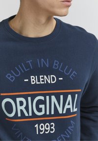 Marineblauwe sweatshirts met een ronde halslijn, voorzien van opvallende witte en oranje tekst: "BUILT IN BLUE", "BLEND" en "ORIGINAL 1993." Zachte stoftextuur.