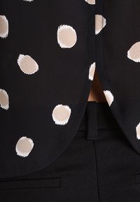 Blusa nera con grandi pois color crema. Presenta una texture liscia e un orlo curvo, abbinata a pantaloni neri su misura.