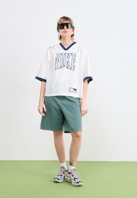 Bílý síťovaný dres Nike s námořnickými akcenty, zelené denimové šortky a šedé tenisky s černými a zelenými detaily, na světlém pozadí.