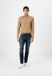 Beige turtleneck-paita, slim fit tummat farkut ja valkoiset lenkkarit. Malli seisoo rentona yksinkertaista taustaa vasten.