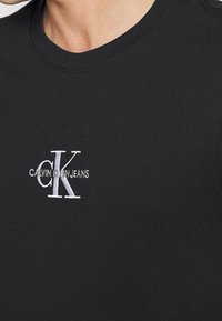 Camiseta de algodón negra con cuello redondo y mangas cortas, con el logo bordado en blanco "CK" y el texto "CALVIN KLEIN JEANS".