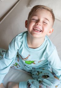 Ljusblå långärmad pyjamasset med lekfulla djurillustrationer, mjukt tyg och avslappnad passform, med ett leende barn sittande på en yta.