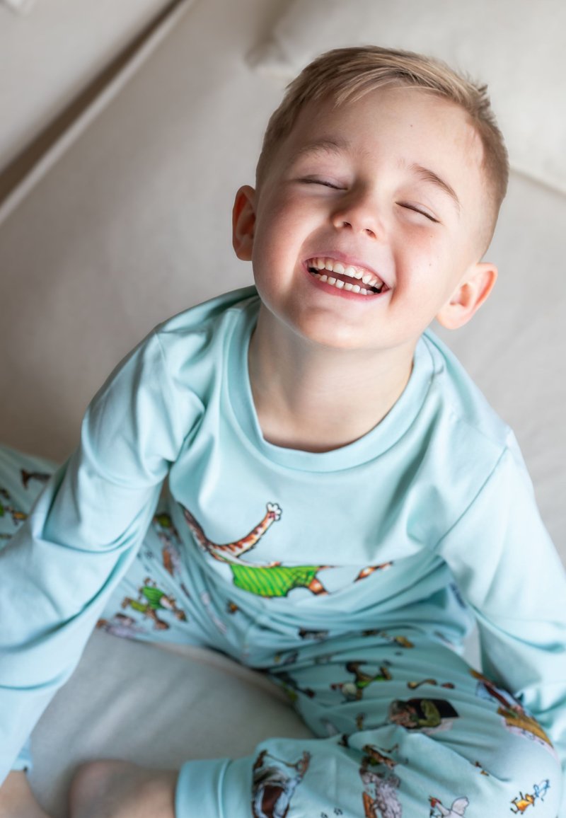 Ljusblå långärmad pyjamasset med lekfulla djurillustrationer, mjukt tyg och avslappnad passform, med ett leende barn sittande på en yta.