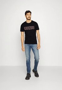 Black cotton T-shirt with "GUESS" logo in red text, paired with light blue jeans and black lace-up shoes, standing in a neutral background.