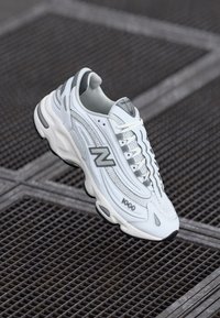 Hvide og grå New Balance 1000 sneakers med snørebånd, mesh-paneler og polstret sål på metalrist baggrund.