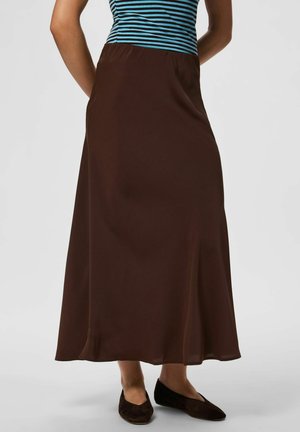 Personne portant une jupe marron longue jusqu'aux chevilles, associée à des chaussures plates noires et un haut rayé bleu et noir, debout, les mains dans le dos.