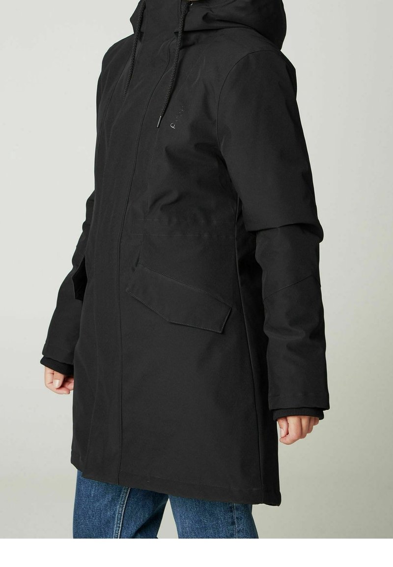 Person wearing a black hooded coat with front flap pockets and blue jeans, standing against a light neutral background.