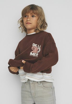 Šviesiaplaukis vaikas dėvi rudą megztinį su užrašu „De La Mer“ ir grafiniu piešiniu, vilkintis dryžuotą marškinių ir šviesių džinsų sluoksnį.