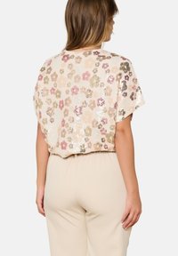 Donna vista di spalle che indossa un top floreale a maniche corte con paillettes nei toni del rosa chiaro, verde e beige, abbinato a pantaloni color crema.