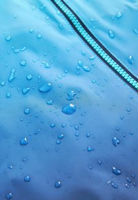 Des gouttes d'eau perlent sur un tissu imperméable bleu vif près d'une fermeture éclair turquoise fermée, indiquant une résistance à l'eau.