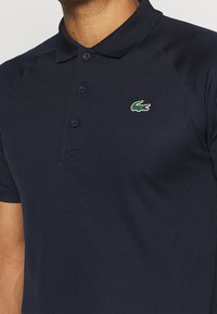 Marineblaues Poloshirt mit Kragen, Knopfleiste, strukturiertem Stoff und kleinem grünen Krokodil-Logo auf der linken Brust.