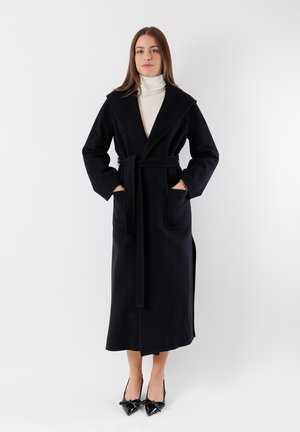 Cappotto avvolgente nero in lana con un ampio colletto, cintura e tasche laterali; di lunghezza fino al ginocchio con una texture liscia. Indossato sopra un dolcevita crema.