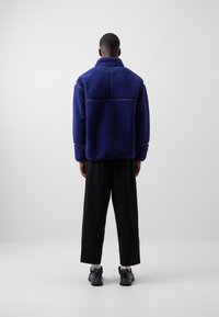 Mørkeblå fleecejakke med høj krave, struktureret stof og sidesømme. Bæres med sorte bukser og sko. Set bagfra.