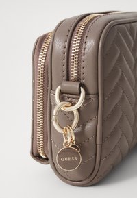 Borsa in tessuto trapuntato marrone con cerniere e accessori dorati. Presenta un'etichetta circolare con il logo "GUESS" e un accento in catena.