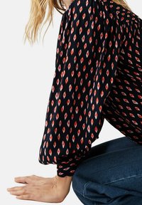 Blusa nera con motivi a diamante rossi e bianchi, maniche lunghe e a sbuffo, e una texture morbida. Abbinata a jeans in denim blu.