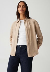 Vrouw in een beige blouse met knopen over een witte top, gecombineerd met donkerblauwe jeans, staande tegen een effen lichte achtergrond.