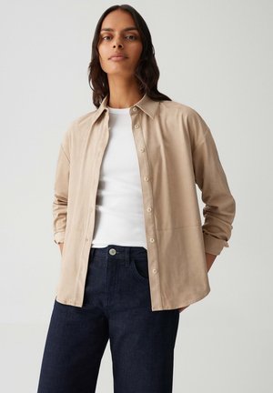 Vrouw in een beige blouse met knopen over een witte top, gecombineerd met donkerblauwe jeans, staande tegen een effen lichte achtergrond.