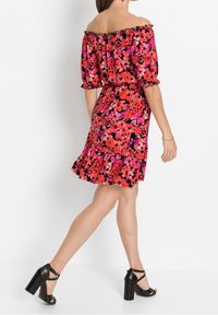 Červené květinové šaty s odhalenými rameny, nafouklými rukávy, zdůrazněným pasem a volánkovým lemem. Nošeny s černými sandály na vysokém podpatku.