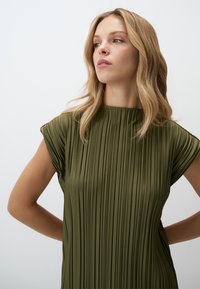 Abito plissettato verde oliva con scollatura alta e maniche a cappuccio. Il tessuto ha una consistenza morbida e liscia, che esalta il design strutturato.