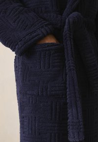 Granatowy pluszowy szlafrok z teksturowanym wzorem, z paskiem i kieszenią po bokach. Tkanina wydaje się miękka i gruba, idealna na ciepło.