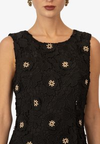 Schwarzes Blumenspitzenkleid mit geschichteten, geprägten Blumen und elfenbeinfarbenen Perlenakzenten. Ärmellos mit rundem Ausschnitt und texturiertem Stoff.